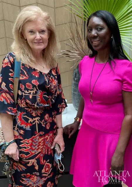 Two women posing for a photo. The woman on the left has blonde hair and wears a dark blue dress with a red and tan pattern. The woman on the right has dark hair and wears a bright pink dress.