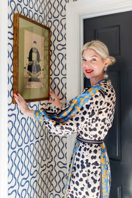 Woman in a leopard print dress stands next to a doorway, touching a framed artwork on a wall with blue and white wavy pattern wallpaper.