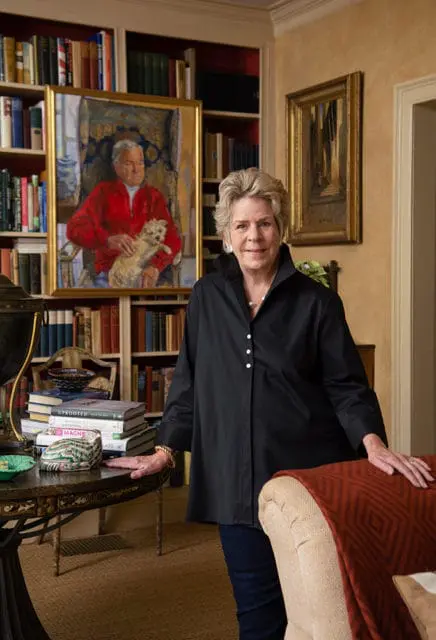Woman in a black collared shirt and jeans stands in a room with bookshelves, a painting of a man and dog, and a draped throw on a chair.