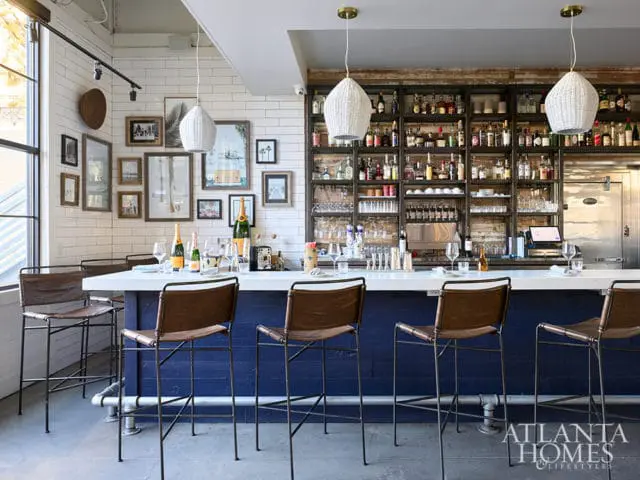 Bar with white countertop, blue base, leather stools, and shelves stocked with liquor bottles.