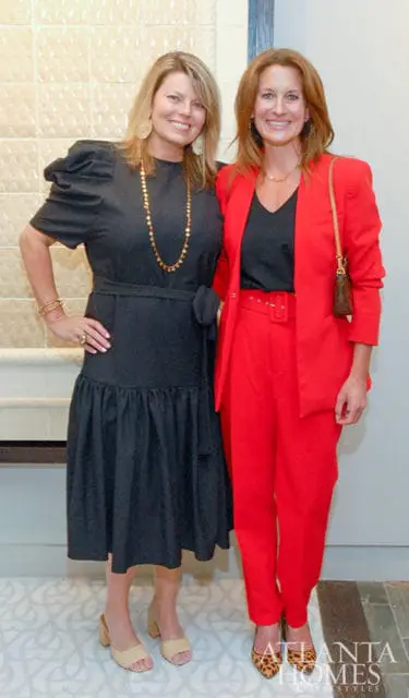 Two women smiling, one in a black dress and gold necklace, the other in a red suit.