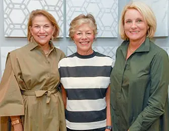 Three women smiling at the camera, standing in front of a wall with geometric tile patterns.