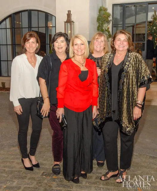 Five women pose for a photo outdoors in front of a building with large windows and a Christmas tree visible in the background. The woman in the center wears a bright red top and black skirt.