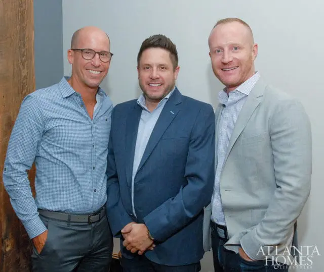 Three men smiling, one in a blue patterned shirt and gray pants, one in a blue suit, and one in a