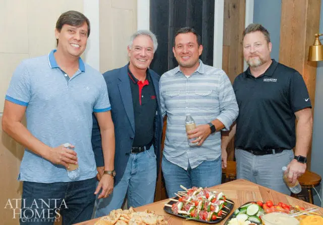 Four men standing together. One wears a blue polo, another a navy blazer over a red-trimmed shirt