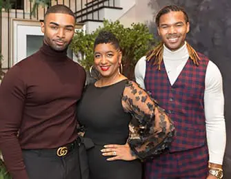 Three people posing for a photo indoors. The person on the left wears a maroon turtleneck. The