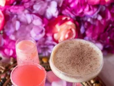 Two cocktails on a mirrored tray with rose petals and a green leaf.
