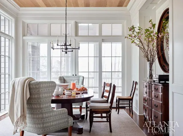 Dining room with round table, upholstered chairs, wood ceiling, and large windows.