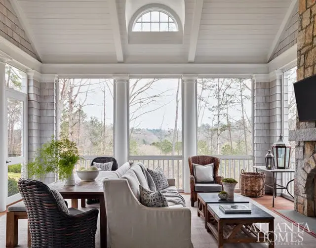 Screened porch with stone fireplace, wicker chairs, and large windows overlooking trees.