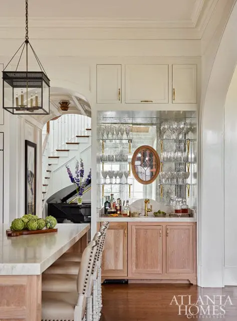 Bar with mirrored backsplash, glass shelves, and wood cabinetry. Piano and staircase visible.
