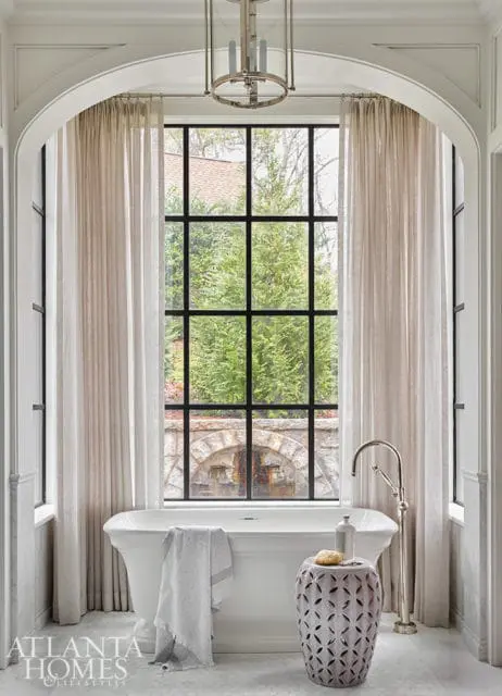 Freestanding white bathtub with a towel, next to a decorative stool and a tall faucet, in front of