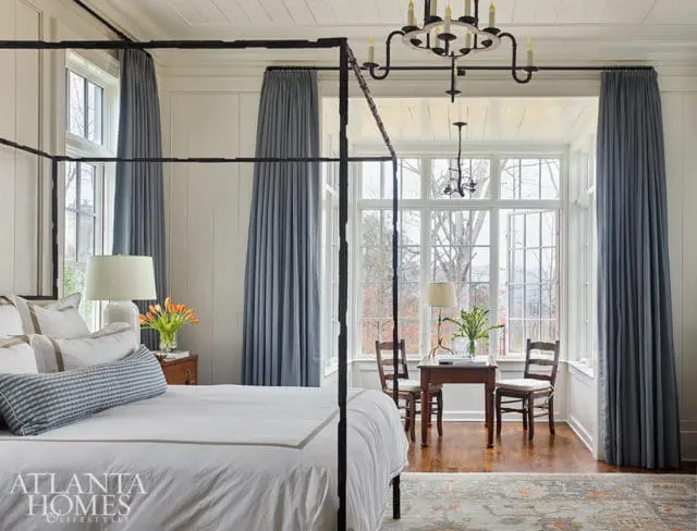 Bedroom with four-poster bed, blue curtains, and bay window overlooking trees.