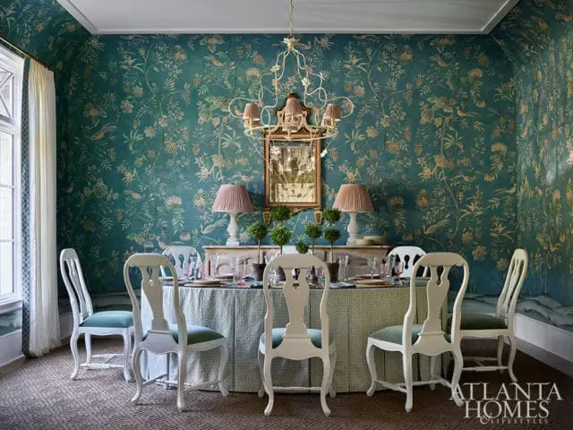 Dining room with teal floral wallpaper, white chairs with green seats, and a patterned tablecloth.