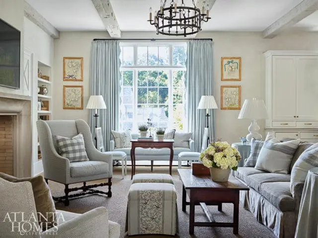Living room with wingback chairs, sofa, and floral arrangements. Light blue curtains frame a large
