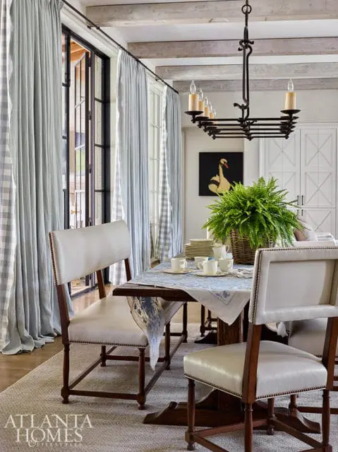 Dining room with light blue gingham curtains, a dark wood table set with white dishes and a fern centerpiece, and upholstered chairs.