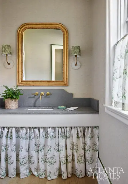 Powder room with gray stone vanity, gold mirror, and floral skirted cabinet.