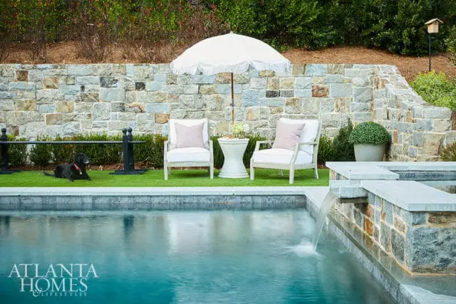 Stone wall with two white chairs, umbrella, and black dog by pool