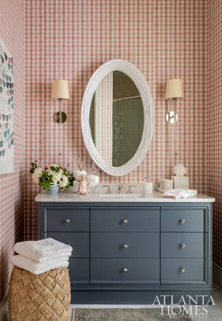 Bathroom with pink gingham wallpaper, gray vanity, white oval mirror, and sconce lighting.