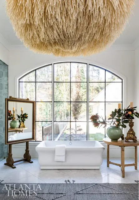 Freestanding bathtub under a large arched window with a textured chandelier above.