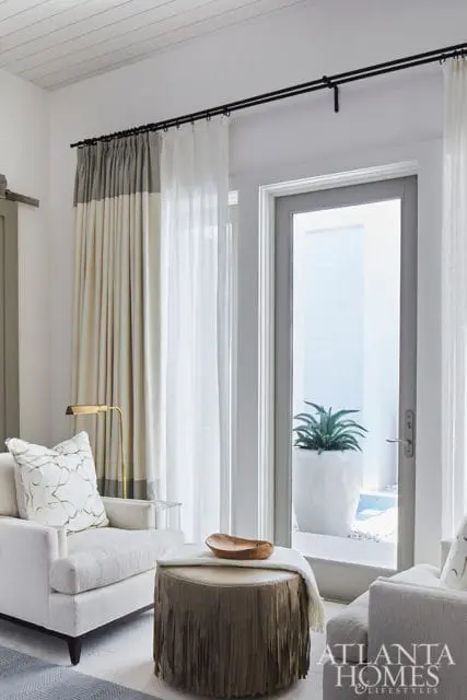 Living room with white armchair, fringed ottoman, and floor-to-ceiling curtains.