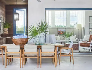 Dining table with wooden chairs, basket of towels, and palm fronds. Living area beyond.