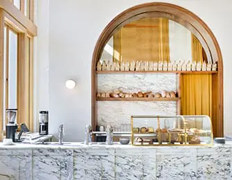 Marble counter with coffee grinders, sink, and display cases filled with baked goods under a wooden
