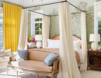 Bedroom with a four-poster bed, floral wallpaper, yellow curtains, and a camel-colored sofa.