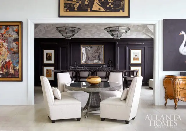 Dining area with light upholstered chairs around a round metal table, leading to a dark bar area.