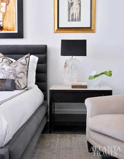 Bedroom with gray upholstered bed, marble-top nightstand with crystal lamp, and beige armchair.