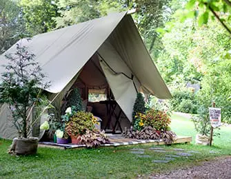 Canvas glamping tent with potted plants and flowers on a wooden deck, surrounded by trees.