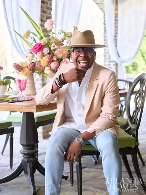 Man in tan hat and peach blazer sitting at a table with floral arrangement.