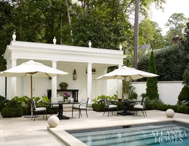 White brick pool house with columns, outdoor dining sets, umbrellas, and pool.