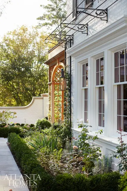 White brick house with arched wooden trellis, wrought iron awning, and manicured garden