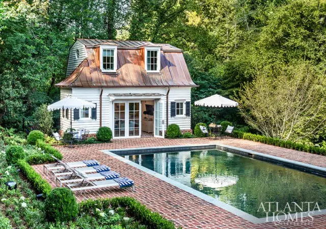 White pool house with copper roof and French doors, brick patio, pool, lounge chairs, and umbrellas.