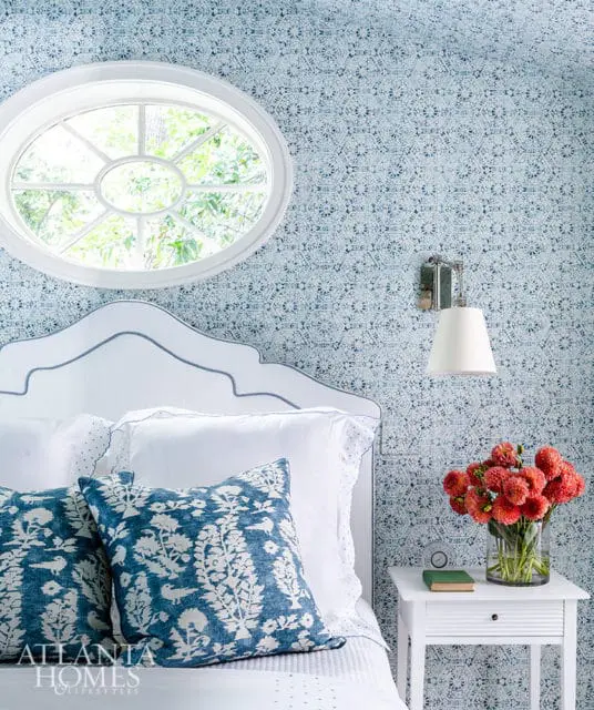 Bedroom with blue patterned wallpaper, white headboard, and blue floral pillows. Oval window.