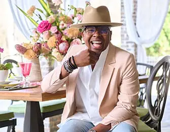 Man in tan fedora and peach blazer smiles at a table set with flowers.