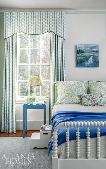 Bedroom with white spindle bed, blue blanket, floral pillows, and patterned curtains.