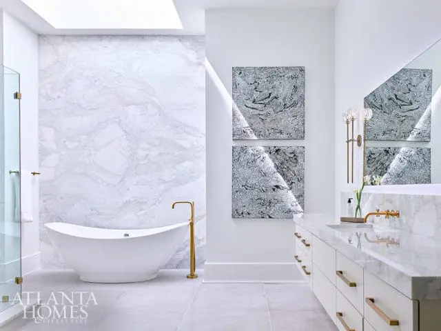 White bathroom with marble accent wall, freestanding tub, and double vanity with brass hardware.