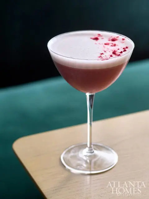 Pink cocktail with foam topping and red garnish in a coupe glass on a wooden table.