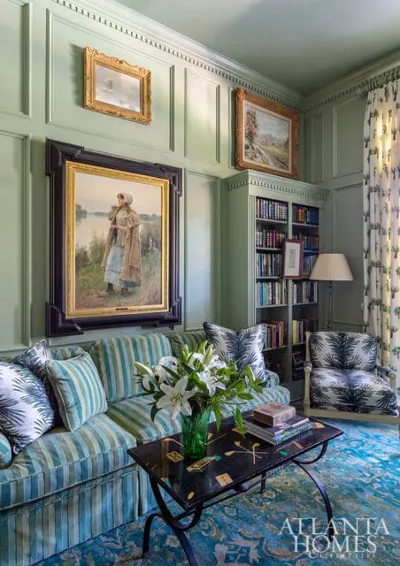 Green paneled living room with striped sofa, armchair, bookshelf, and artwork.