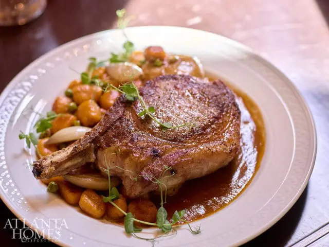 Pork chop with gnocchi, peas, and sauce on a white plate with Atlanta Homes & Lifestyles logo.