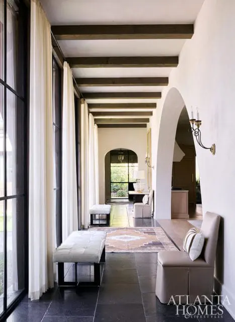 Hallway with dark wood beams, arched doorways, and large windows with white curtains.