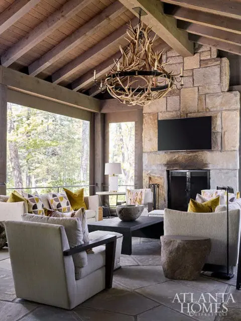 Living room with stone fireplace, antler chandelier, and large windows overlooking trees.
