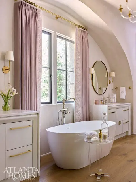 Freestanding white bathtub with chrome faucet, next to a vanity with marble countertop and gold
