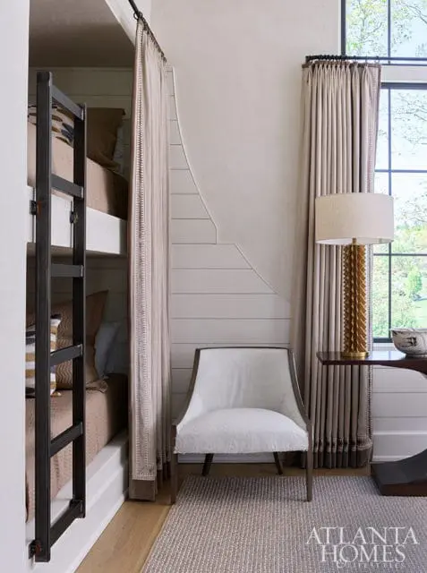 Bunk beds with neutral bedding, white armchair, and gold lamp next to window.