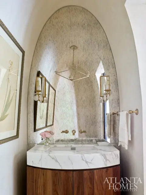 Powder room with marble vanity, wood cabinetry, arched stone wall, and brass fixtures.