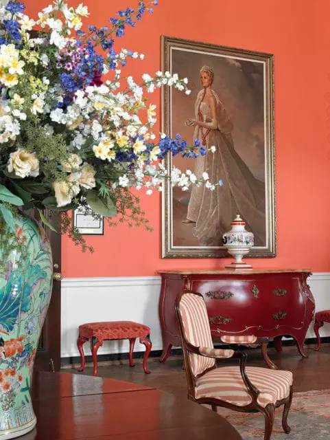 Coral walls with a portrait, red dresser, striped armchair, and floral arrangement