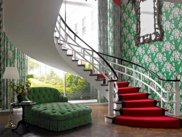 Green tufted chaise lounge, red carpeted spiral staircase, and floral wallpaper