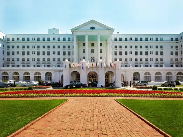 White hotel with columns and arched entrances, lined with red flowers and green grass.
