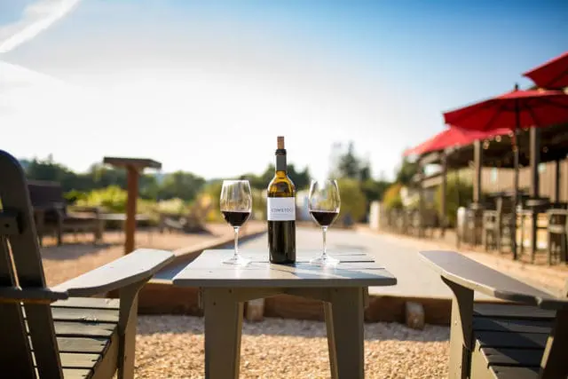 Comstock wine bottle and two glasses on a table with outdoor seating and red umbrellas